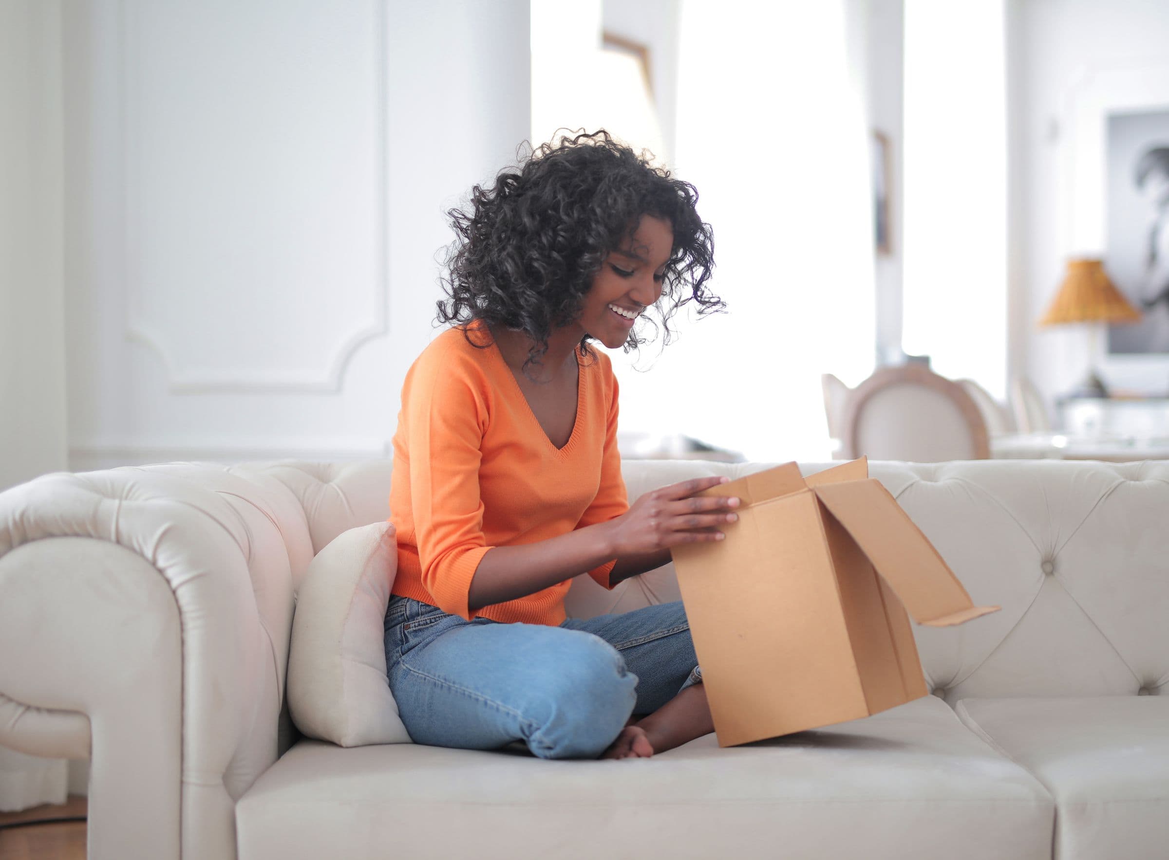A woman on a white sofa at home, smiling as she opens a delivery box